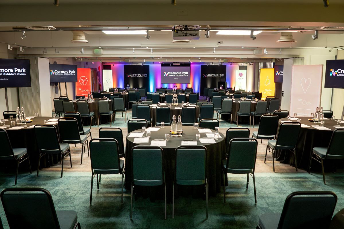 A spacious conference room set up for an event, featuring rows of round tables with black tablecloths and chairs. Colorful banners and screens are displayed at the front, with soft lighting enhancing the atmosphere.